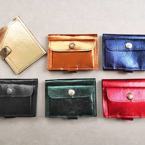 May include: A selection of metallic wallets in gold, copper, blue, black, green, and red. Each wallet has a small pocket with a snap closure. The wallets are displayed on a neutral grey surface.