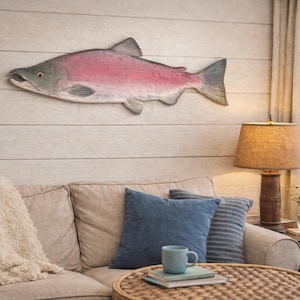 May include: A wall-mounted wooden fish carving featuring a pink and grey body with detailed scales and fins. The fish is positioned horizontally with its head facing left, showcasing a realistic eye and open mouth design on a light-colored wall.