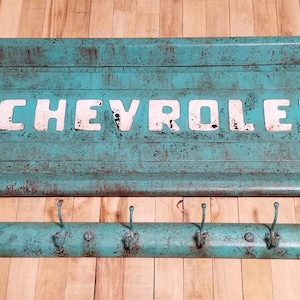 May include: A turquoise blue metal sign with the word "CHEVROLET" in white letters. The sign is distressed and has a rusty patina. It is attached to a turquoise blue metal coat rack with five hooks.