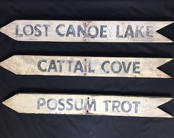 May include: Three wooden signs with arrowheads point to different locations. The signs are painted white and have black lettering. The top sign reads "Lost Canoe Lake", the middle sign reads "Cattail Cove", and the bottom sign reads "Possum Trot".