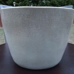 Puede incluir: Una maceta grande de cerámica blanquecina con acabado craquelado. La maceta tiene una forma redondeada y cónica. Se encuentra sobre una superficie oscura. El fondo incluye una valla de madera y hierba verde.