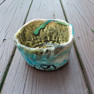 May include: A small, handmade ceramic bowl with a unique, abstract design. The bowl is glazed in shades of blue, green, and yellow, with a textured surface.