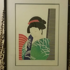 Op de afbeelding: Ingelijste prent van een geisha in een groene kimono met witte accenten en een zwart kapsel. Het kunstwerk heeft een rood-wit gestreept ontwerp en een zwart-wit gestreept verticaal element. De lijst is zwart.