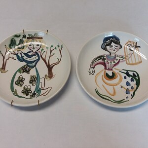 May include: Two white ceramic plates with hand-painted designs of women in colorful dresses. One plate features a woman holding a bird in a cage, while the other plate features a woman holding a pear.