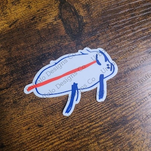 May include: A white sticker with a blue outline of a pig. The pig has a red line across its body and the text "Buffalo Designs Co" is written on the sticker.