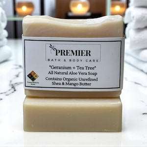 May include: Two bars of Premier Bath & Body Care soap. The top bar is rectangular with rounded edges and a white label that reads "Geranium + Tea Tree" and "All Natural Aloe Vera Soap." The soap contains organic shea and mango butter.