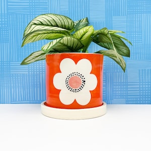 May include: Orange ceramic planter with a white flower design. The flower has a pink center and black dots. The planter has a white rim and a white saucer.