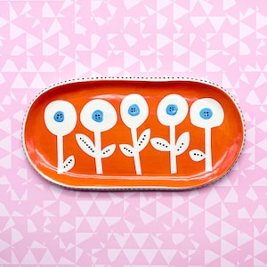 May include: Orange ceramic oval dish with a white rim and black dots. The dish is decorated with five white flowers with blue centers and white stems.