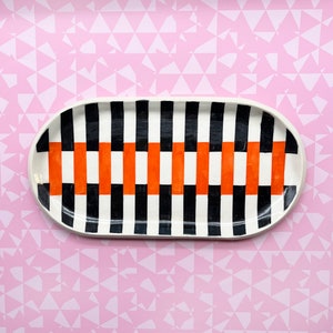 May include: An oval ceramic serving platter with a bold geometric design. The pattern features vertical black and white stripes, interspersed with orange rectangular blocks. The platter has a white rim and is set against a pink background.