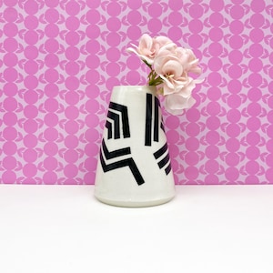 May include: A white ceramic vase with black geometric designs holds pink flowers. The vase has a flared shape and is set against a pink patterned background. The flowers are a soft pink color.