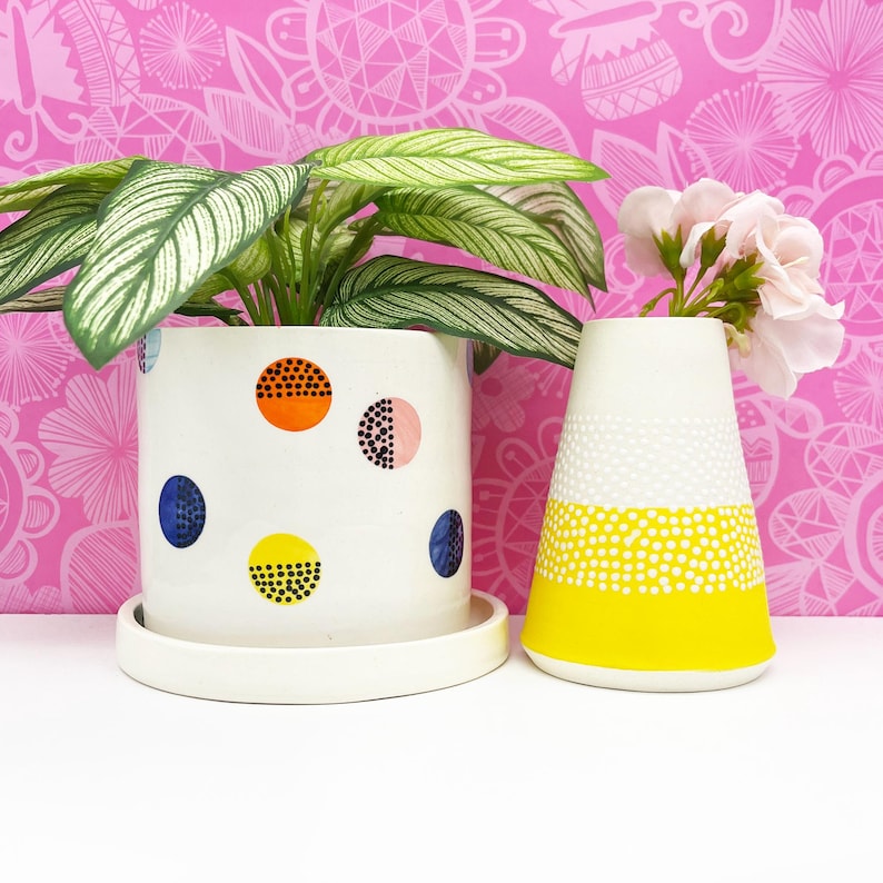 Pode incluir: Dois vasos de cer&acirc;mica branca com padr&otilde;es de bolinhas coloridas. Um vaso &eacute; um cilindro com um pires e cont&eacute;m uma planta verde. O outro vaso &eacute; em forma de cone e cont&eacute;m flores cor-de-rosa.