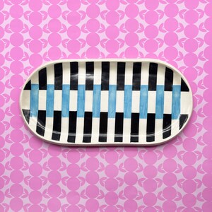 May include: An oval ceramic serving platter with a geometric pattern of black, white, and light blue vertical stripes. The platter has a white rim and is set against a pink background with a repeating pattern of circles and leaves.