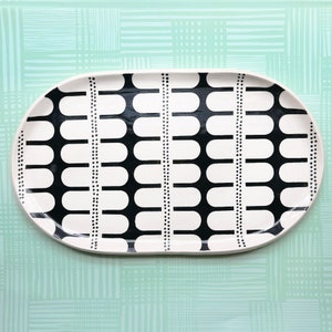 May include: An oval serving platter with a black and white geometric pattern. The white surface is decorated with vertical rows of black, rounded shapes and dotted lines. The platter is on a light green background.