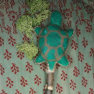 May include: A handmade turtle-shaped staff featuring a green shell and a wooden handle. The turtle has brown detailing and is attached to a long, brown wooden pole. Green moss is attached to the top of the staff. The staff is set against a patterned fabric.