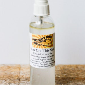 Puede incluir: Una botella de spray de vidrio transparente con una tapa blanca y una etiqueta que dice "Dark Dirty Hippie You Got This Shit!". La etiqueta describe el contenido como un spray de aceite esencial para manifestar y obtener energía. La botella está sobre una superficie de madera.