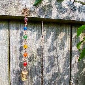 May include: A handmade wind chime with a copper hook and chain, featuring colorful glass beads in a rainbow pattern. The chime hangs from a weathered wooden fence and ends with a small, gold-colored bell.