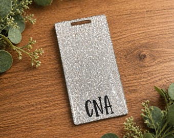 May include: A rectangular, silver glitter badge holder with a hole at the top. The letters "CNA" are printed in black on the lower portion of the badge holder. The badge holder is on a wooden surface, surrounded by green foliage.