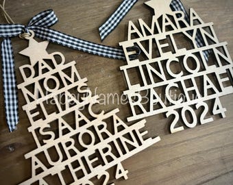 Puede incluir: Dos adornos de árbol de Navidad de madera con nombres y el año 2024 recortados. Los adornos son de madera de color claro y tienen una estrella en la parte superior. Cada adorno está adornado con una cinta a cuadros en blanco y negro.