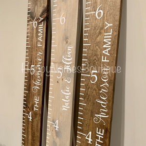May include: Three wooden growth charts with white painted numbers and names. The charts are labelled "The Hammer Family", "Natalie & Jillian", and "The Anderson Family".