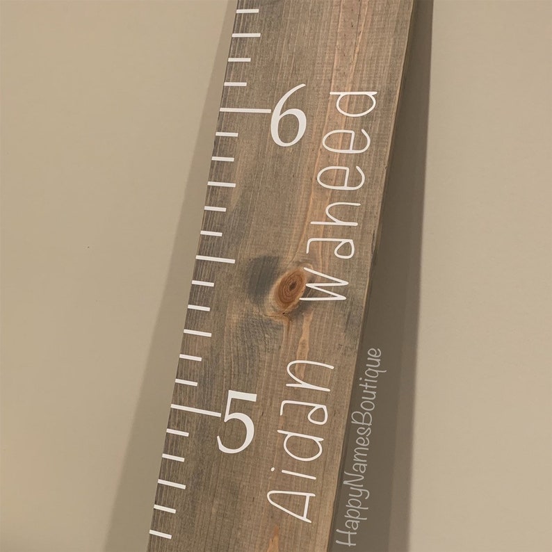 May include: A wooden growth chart with white numbers and the name "Aidan Waheed" written on it. The chart is made of rustic wood and has a distressed look. The text "HappyNamesBoutique" is printed at the bottom of the chart.