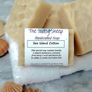 May include: Beige handcrafted soap bars with a textured surface, wrapped in a white paper label reading The Sudsy Sheep Handcrafted Soap Sea Island Cotton, accompanied by small tan seashell-shaped soap pieces on a white fabric square.