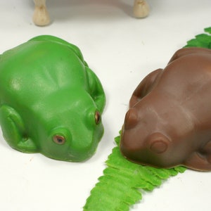 May include: Two frog-shaped soaps, one green and one brown, on a green leafy surface.