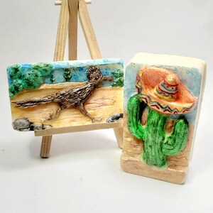 May include: Two rectangular soap bars with raised relief designs. One bar features a brown roadrunner in a desert landscape on a wooden easel. The second bar displays a green cactus wearing a colorful sombrero against a blue sky background.