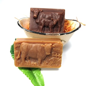 May include: Two bars of soap with cow designs. One bar is brown and features a cow and calf. The other bar is tan and features a single cow. Both bars are in a white ceramic bowl with straw-like material.