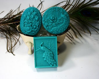 May include: Three teal blue soap bars with raised peacock designs. The soaps are displayed in a white ceramic bowl with three legs.