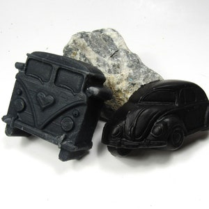 May include: Black soapstone carvings of a vintage VW bus with a heart design and a classic Beetle car. A gray rock is behind the carvings. The objects are on a white surface.
