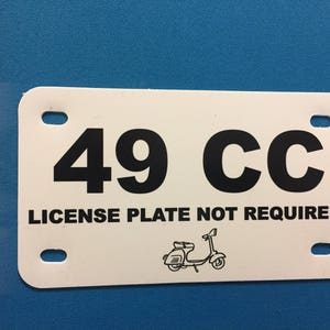 May include: White rectangular sign with black text that reads "49 CC LICENSE PLATE NOT REQUIRED". A small black and white illustration of a scooter is in the bottom center of the sign.
