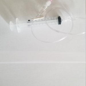 May include: A clear plastic syringe with a black tip and a white plastic connector. A clear plastic tube is attached to the syringe and a pink plastic tube is connected to the connector.