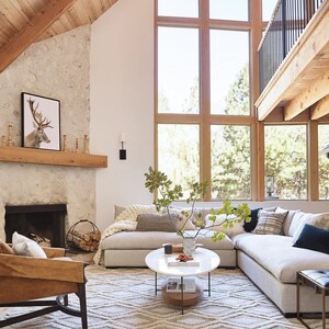 Pode incluir: Um grande sof&aacute; seccional com estofamento branco e uma variedade de almofadas decorativas fica em uma sala de estar com uma lareira de pedra e uma grande janela. Uma mesa de centro com tampo de m&aacute;rmore branco fica na frente do sof&aacute;. Um tapete com um padr&atilde;o geom&eacute;trico cobre o ch&atilde;o.