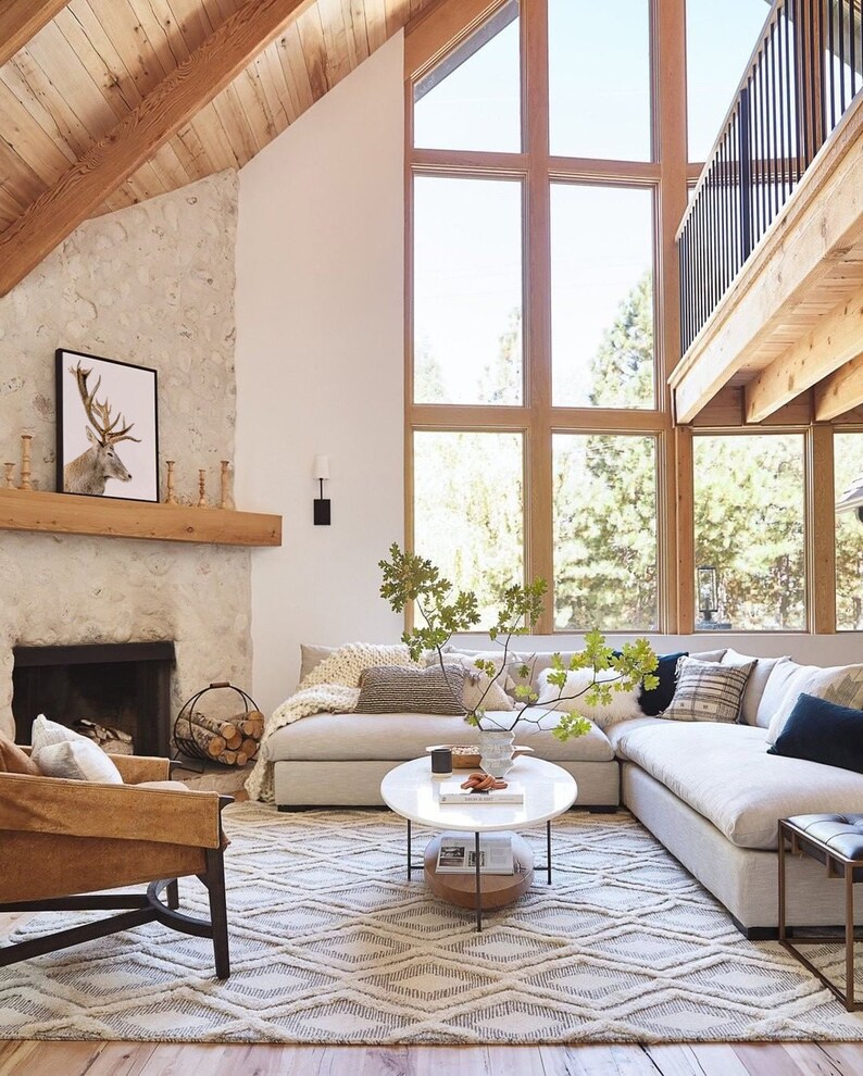Pode incluir: Um grande sof&aacute; seccional com estofamento branco e uma variedade de almofadas decorativas fica em uma sala de estar com uma lareira de pedra e uma grande janela. Uma mesa de centro com tampo de m&aacute;rmore branco fica na frente do sof&aacute;. Um tapete com um padr&atilde;o geom&eacute;trico cobre o ch&atilde;o.