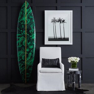 May include: A white armchair with a black pillow sits in front of a black and green surfboard. A black and white photo of palm trees hangs on the wall behind the chair. A small black and white marble side table with a vase of white flowers sits to the right of the chair.