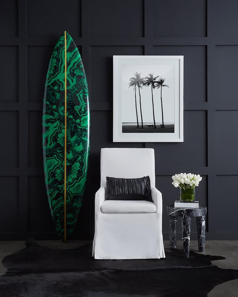 May include: A white armchair with a black pillow sits in front of a black and green surfboard. A black and white photo of palm trees hangs on the wall behind the chair. A small black and white marble side table with a vase of white flowers sits to the right of the chair.