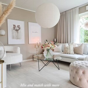K&ouml;nnte beinhalten: Ein Wohnzimmer-Interieur mit einem wei&szlig;en, flauschigen Sessel, einem wei&szlig;en Sofa mit Kissen, einem Glas-Couchtisch mit schwarzem Metallgestell, einer wei&szlig;en runden Pendelleuchte und einem rosa gepolsterten Hocker.