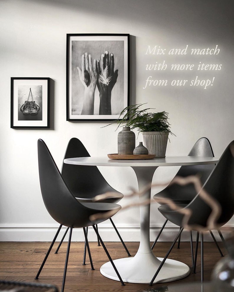 May include: A white round table with a black tulip base and four black chairs. The table is set with a wooden tray, two small gray vases, and a larger gray planter with a green plant. Two black and white framed prints hang on the wall behind the table. The text "Mix and match with more items from our shop!" is written on the wall.