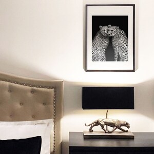 May include: A black and white photograph of two cheetahs cuddling, framed in a black frame and hanging on a white wall above a nightstand with a lamp and a gold cheetah statue.
