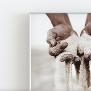 K&ouml;nnte beinhalten: Nahaufnahme von zwei H&auml;nden, die zusammengelegt sind und Sand durch die Finger rieseln lassen. Der Sand ist hellbeige und die H&auml;nde sind braun.