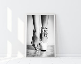May include: Black and white photo of a ballerina's feet in pointe shoes. The ballerina is standing on her toes, and the photo is taken from a low angle. The photo is in a white frame.