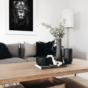 May include: A black and white photo of a lion's face in a white frame hangs above a wooden coffee table. The table has a woven basket, a book, a black bowl, and a string of white beads on it. A vase with eucalyptus branches sits on the table.