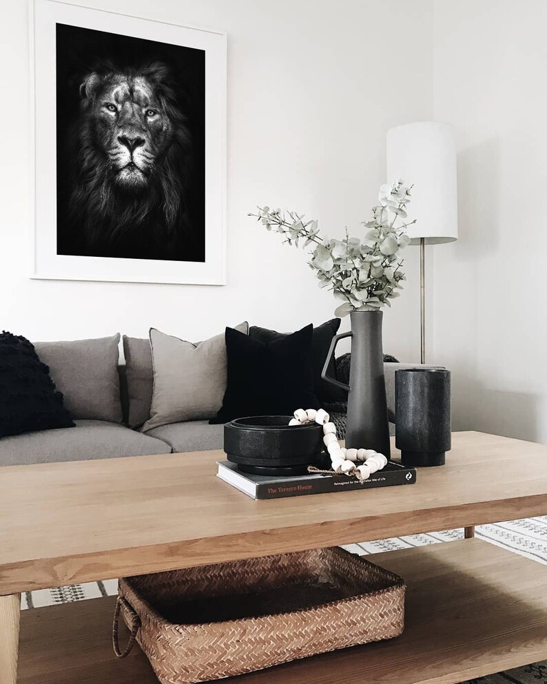 May include: A black and white photo of a lion's face in a white frame hangs above a wooden coffee table. The table has a woven basket, a book, a black bowl, and a string of white beads on it. A vase with eucalyptus branches sits on the table.