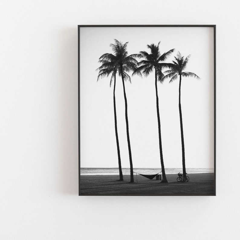 May include: Black and white photograph of four palm trees silhouetted against a white sky and a beach. A hammock hangs between two of the trees and a bicycle is parked on the beach.