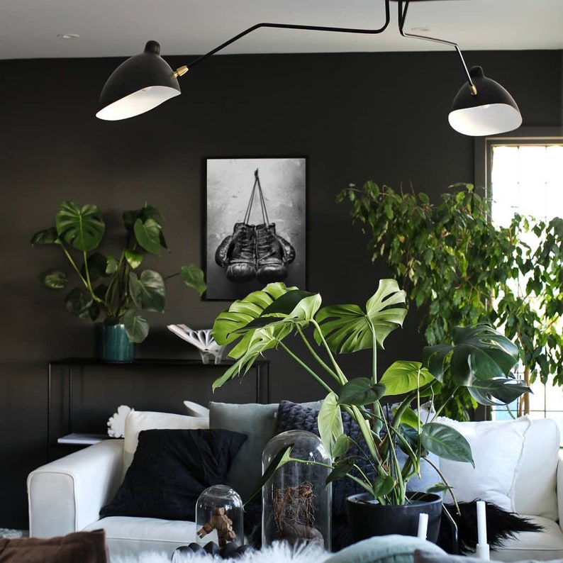 May include: A black and white photograph of a pair of boxing gloves hanging from a hook. The photo is framed and hanging on a dark wall in a living room with plants and a white couch.