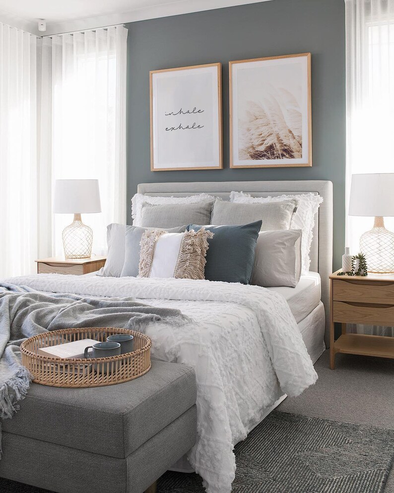 May include: A white bed with a gray headboard and a white duvet cover. The bed is decorated with pillows in various shades of gray, white, and blue. There are two framed prints on the wall behind the bed, one with the words "inhale exhale" and the other with a nature scene. There is a gray ottoman in front of the bed with a woven tray on top.