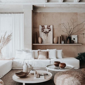 K&ouml;nnte beinhalten: Ein wei&szlig;es Ecksofa mit wei&szlig;en Kissen und einem braunen Zierkissen in einem Wohnzimmer mit einem wei&szlig;en Couchtisch und einem runden gewebten Hocker. Der Couchtisch hat eine Glasplatte und ein wei&szlig;es Gestell. An der Wand hinter dem Sofa h&auml;ngt ein gro&szlig;er Druck. Der Druck zeigt ein Paar H&auml;nde, die Sand halten.