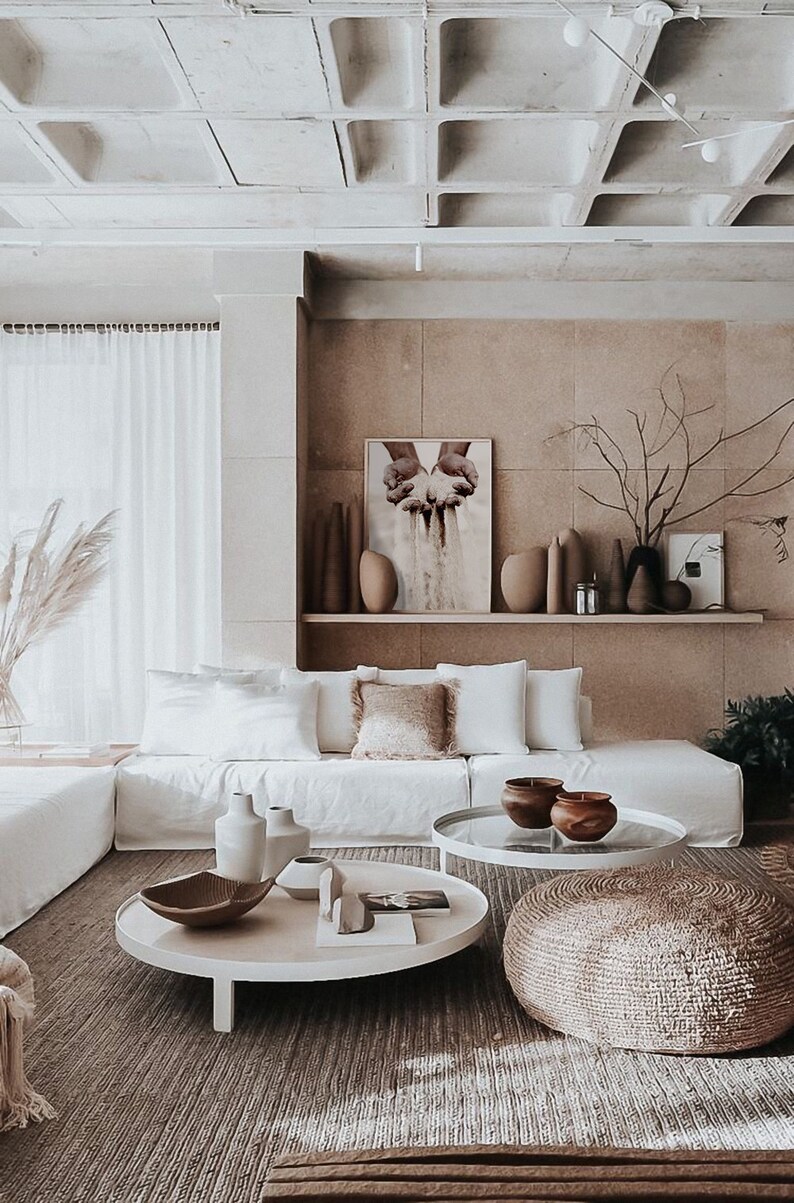K&ouml;nnte beinhalten: Ein wei&szlig;es Ecksofa mit wei&szlig;en Kissen und einem braunen Zierkissen in einem Wohnzimmer mit einem wei&szlig;en Couchtisch und einem runden gewebten Hocker. Der Couchtisch hat eine Glasplatte und ein wei&szlig;es Gestell. An der Wand hinter dem Sofa h&auml;ngt ein gro&szlig;er Druck. Der Druck zeigt ein Paar H&auml;nde, die Sand halten.