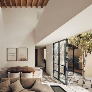 May include: A modern living room with a large, open floor plan. The room features a large, light-colored sofa with pillows, a coffee table, and a dining table with chairs. The room has a large window that looks out onto a patio with a tree and chairs. The walls are white and the ceiling has exposed wooden beams. The artwork on the wall says "inhale exhale".