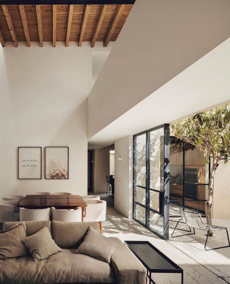 May include: A modern living room with a large, open floor plan. The room features a large, light-colored sofa with pillows, a coffee table, and a dining table with chairs. The room has a large window that looks out onto a patio with a tree and chairs. The walls are white and the ceiling has exposed wooden beams. The artwork on the wall says "inhale exhale".
